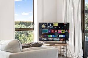 A television mounted on a stand in a room with a large window, a white couch, and a curtain. - Charming single-level home - just moments from the beach,  sun-soaked garden. (Auckland)