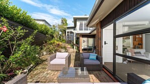 A modern outdoor patio with wicker furniture, a glass door leading to a living area, and a brick pathway. - Charming single-level home - just moments from the beach,  sun-soaked garden. (Auckland)