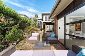 A modern outdoor patio with wicker furniture, a glass door leading to a living area, and a brick pathway. - Charming single-level home - just moments from the beach,  sun-soaked garden. (Auckland)
