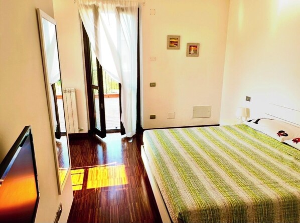 A hotel room with a bed, a television, a window with curtains, and a door leading to a balcony.