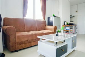 A living room with a brown sofa, a white coffee table, a small plant, and a kitchen area with a kettle and utensils. - Cozy And Simple 1Br At Apartment Grand Sentraland Karawang (Karawang)