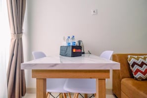 Ein kleiner, moderner Raum mit einem Tisch mit Marmorplatte, zwei weißen Stühlen, einem braunen Sofa mit einem gemusterten Kissen sowie einer Minibar mit Wasserflaschen und einer Kaffeemaschine.