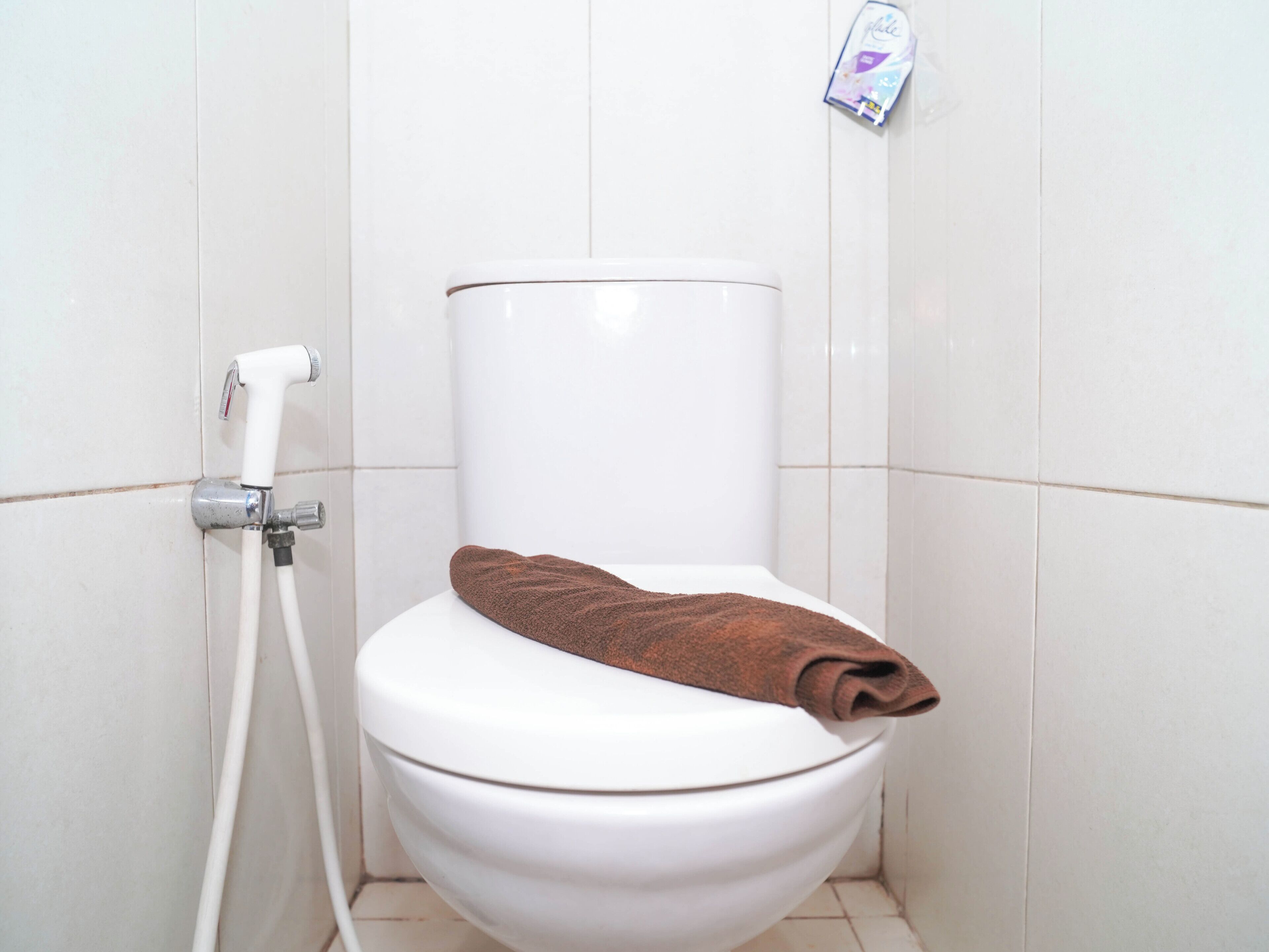 A white toilet with a brown towel on it, a handheld showerhead, and a soap dispenser on the wall.
