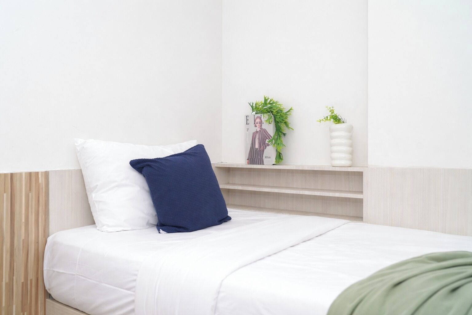 Een bed met witte en marineblauwe kussens, een groene deken en een plank met een plant en een vaas.