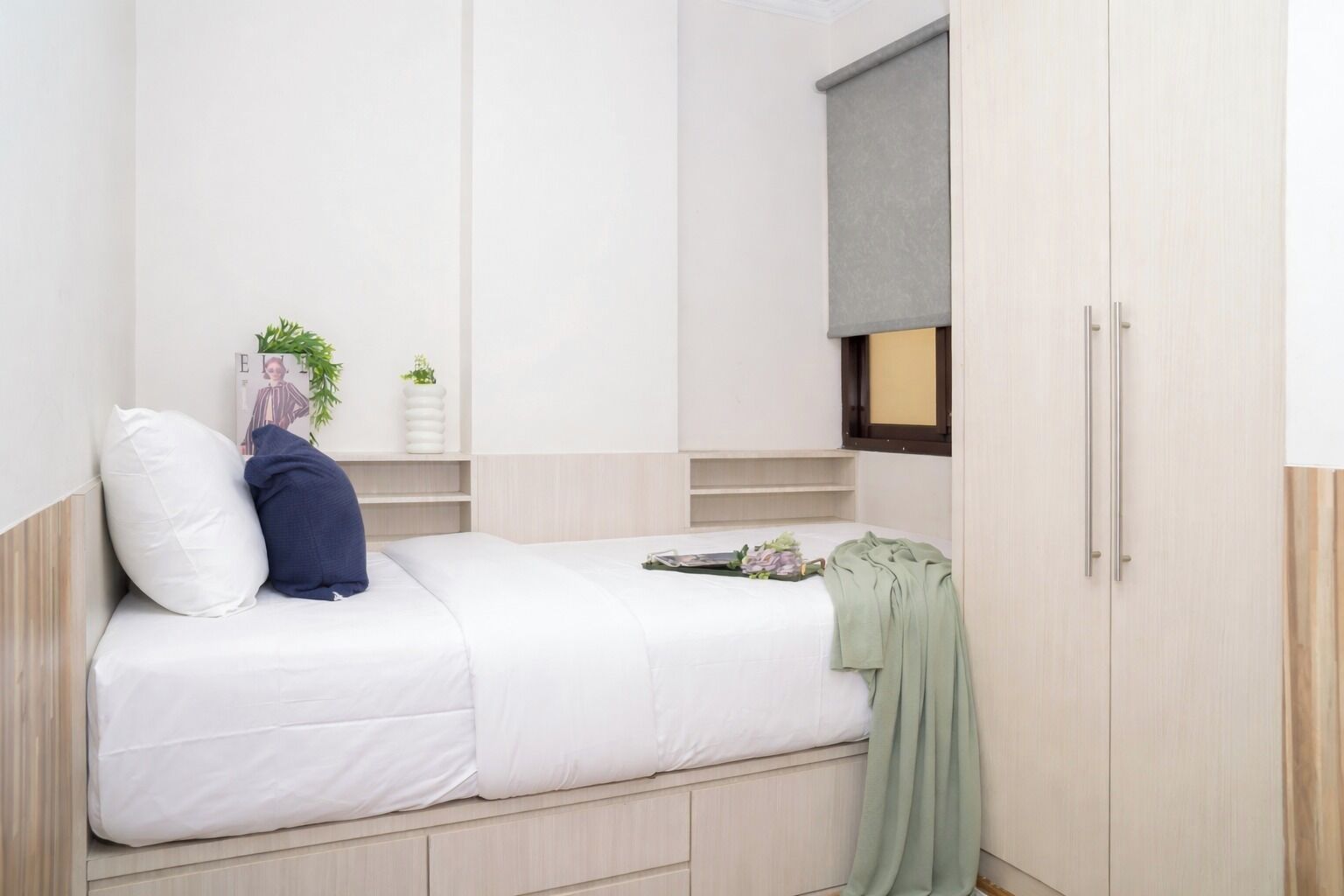A single bed with a white mattress, a blue pillow, and a green throw blanket. There is a small shelf with plants and a magazine. A window with a blind is visible.