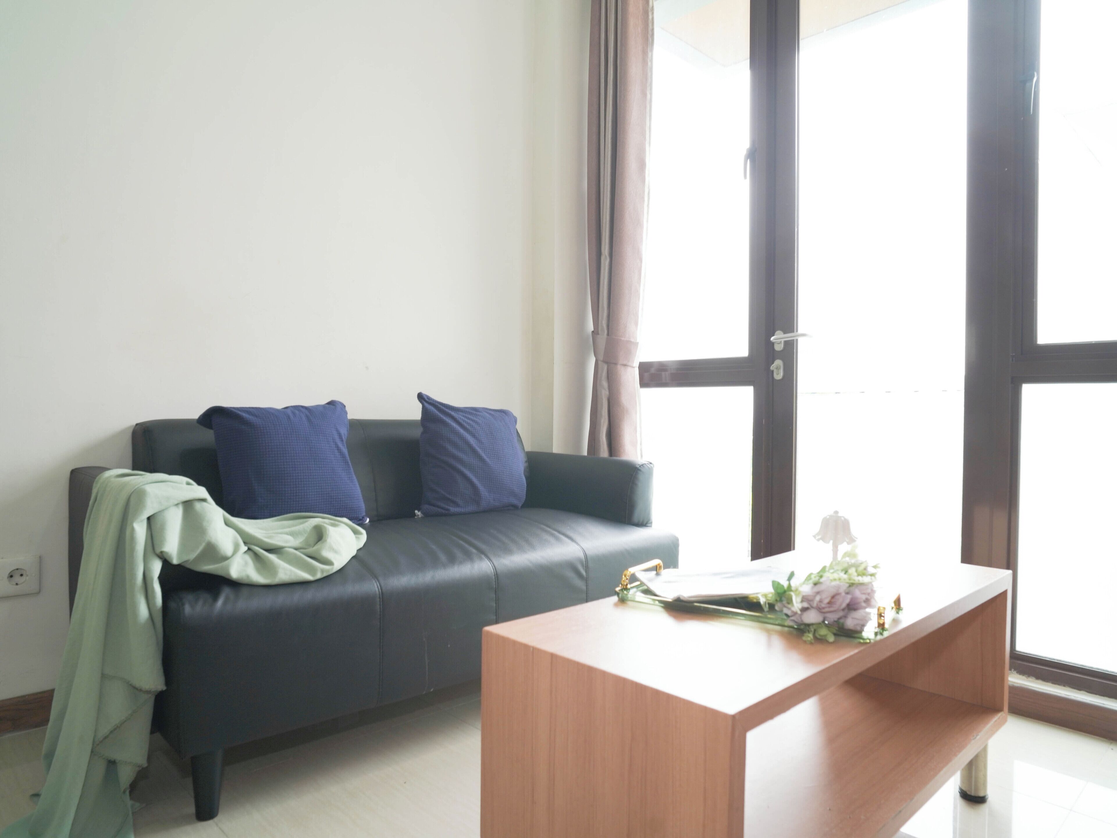 Una sala de estar con un sofá de cuero oscuro, dos cojines azules, un plaid verde y una mesa de centro de madera con un florero.