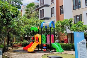 A playground with slides and climbing structures, surrounded by greenery and residential buildings. - Practical 2Br For 3 Pax At Asatti Apartment (Pagedangan)