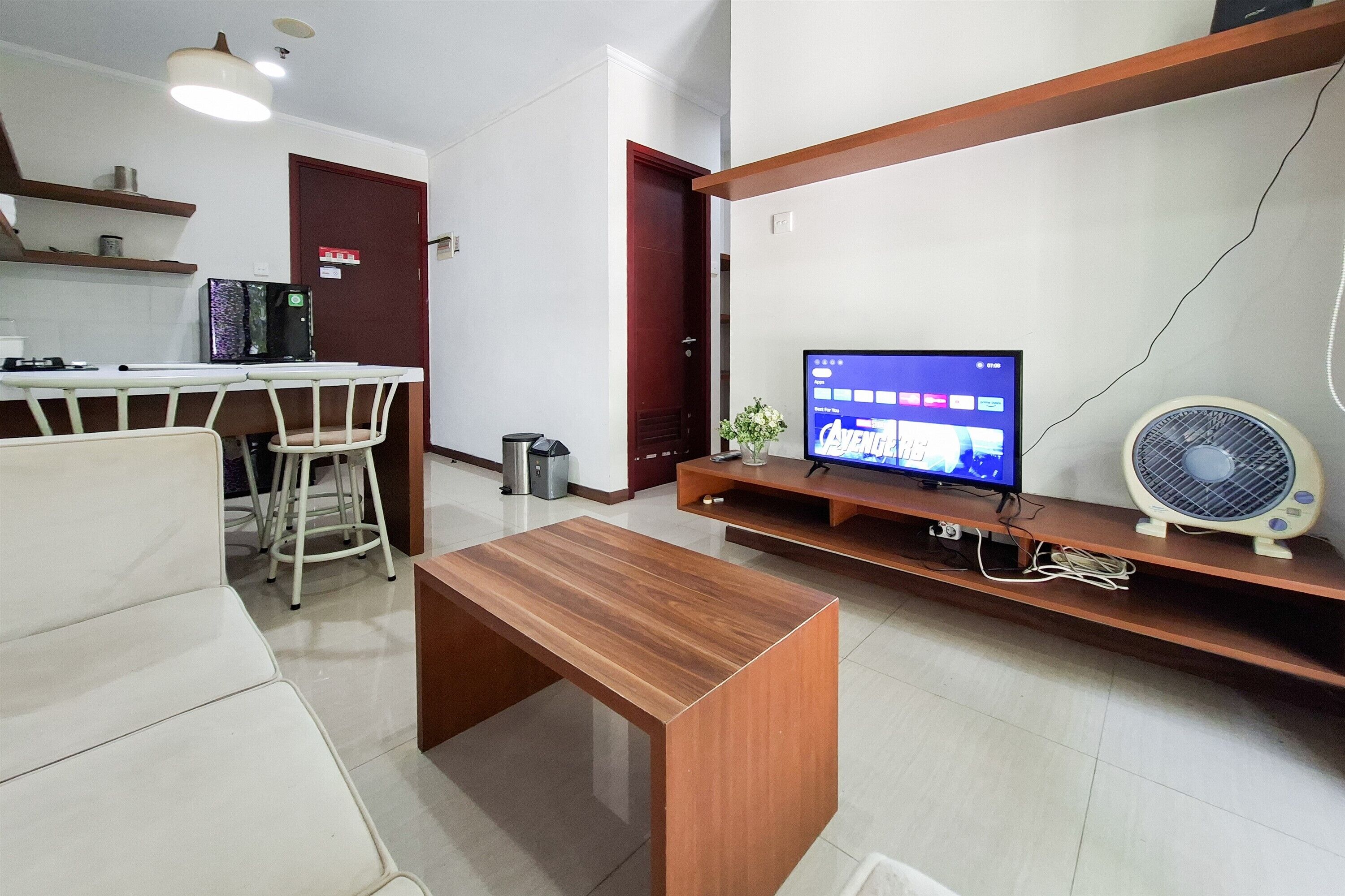 A modern living room with a wooden coffee table, a TV on a stand, a fan, and a kitchen area with a refrigerator and a coffee machine.
