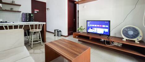 A modern living room with a wooden coffee table, a TV on a stand, a fan, and a kitchen area with a refrigerator and a coffee machine.