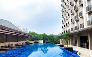 A swimming pool area with lounge chairs and a covered seating area, surrounded by greenery and modern buildings. - Cozy And Roomy Studio Space At Apartment Azalea Suites (Cikarang)
