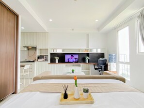 A modern hotel room with a bed, bedside table, TV, kitchenette, and a large window. - Cozy And Roomy Studio Space At Apartment Azalea Suites (Cikarang)