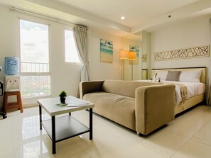 A hotel room with a bed, a sofa, a small table with a plant, a water dispenser, and a window with curtains. - Cozy And Roomy Studio Space At Apartment Azalea Suites (Cikarang)
