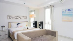 A hotel room with a bed, a sofa, a bedside table, a lamp, a clock, and a painting on the wall. - Clean And Relaxing Studio Room At Apartment Azalea Suites (Cikarang)
