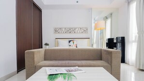 A modern hotel room with a sofa bed, a bedside table with a magazine and a plant, a decorative wall piece, and a water dispenser. - Clean And Relaxing Studio Room At Apartment Azalea Suites (Cikarang)