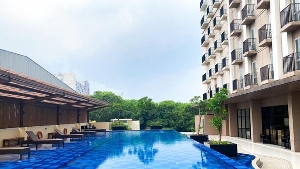 A swimming pool area with lounge chairs and a covered seating area, surrounded by greenery and modern buildings. - Clean And Relaxing Studio Room At Apartment Azalea Suites (Cikarang)