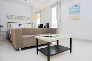 A modern hotel room with a beige sofa, a small table, a bed with white bedding, a bedside table with a lamp, a coffee machine, and a painting of a beach. - Clean And Relaxing Studio Room At Apartment Azalea Suites (Cikarang)