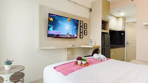 A hotel room with a bed, a TV mounted on the wall, a small table with a plant, and a kitchenette area. - Comfy White Studio For 2 Pax At Bale Hinggil Apartment (Surabaya)