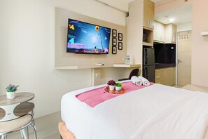 A hotel room with a bed, a TV mounted on the wall, a small table with a plant, and a kitchenette area. - Comfy White Studio For 2 Pax At Bale Hinggil Apartment (Surabaya)