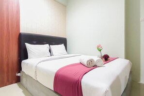 Uma cama bem arrumada com roupa de cama branca e um toque rosa, um pequeno vaso com flores e um relógio na mesa de cabeceira.