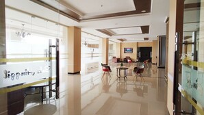Um saguão de hotel com divisória de vidro, balcão de recepção e área de estar com cadeiras e uma mesa.