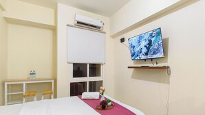 Habitación de hotel con cama, televisión, aire acondicionado, ventana, escritorio con sillas y estante con televisor.