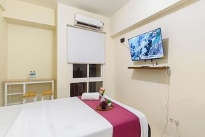 Habitación de hotel con cama, televisión, aire acondicionado, ventana, escritorio con sillas y estante con televisor.