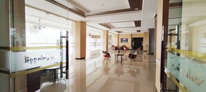 Un hall de hotel con un separador de vidrio, un mostrador de recepción y una zona de estar con sillas y una mesa.