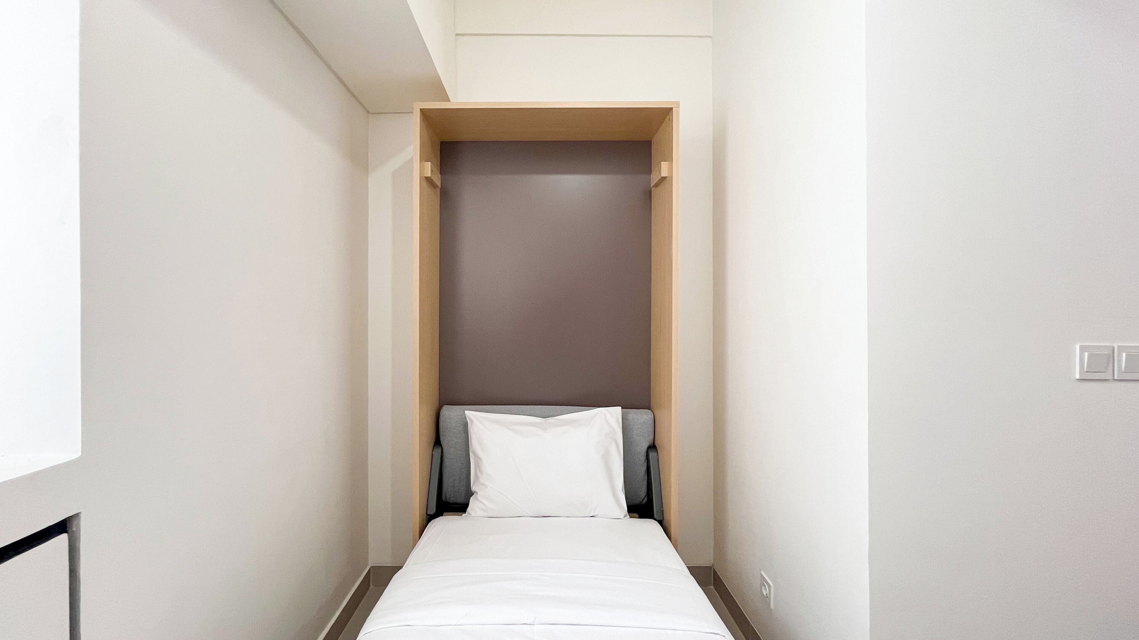 A narrow, single-bed room with a plain white wall and a wooden headboard.