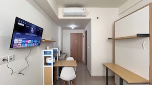 A compact room with a flat-screen TV mounted on the wall, a wooden door, a small table, and a white chair. - Simply Look Studio (No Kitchen) At Bandaraya - Tallasa City Makassar Apartment (Makassar)