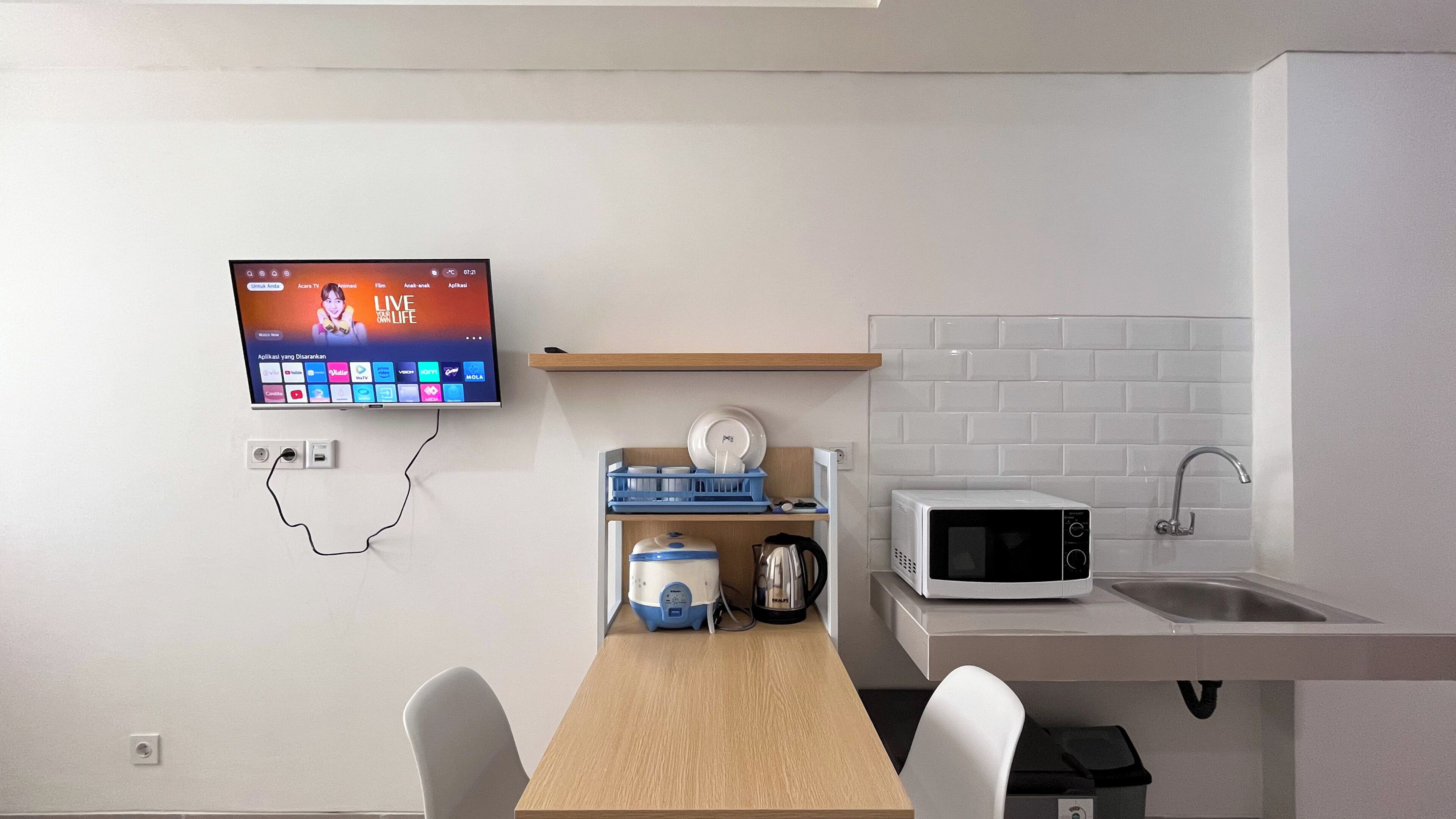 Una cocina moderna con un televisor de pantalla plana en la pared, un microondas, un hervidor y un escurreplatos.