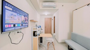 A small, modern room with a flat-screen TV mounted on the wall displaying various streaming services. There's a wooden desk with a chair, a small table, and a light blue sofa. An air conditioning unit is mounted on the wall above the desk. - Warm And Comfy Studio Apartment (No Kitchen) Bandaraya - Tallasa City Makassar (Makassar)