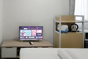 A hotel room with a TV on a wooden desk, a mobile phone, a white bed, and a kitchenette with a kettle and coffee maker. - Best Price Studio (No Kitchen) Apartment At Bandaraya - Tallasa City Makassar (Makassar)