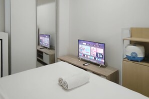 A hotel room with a bed, a TV on a stand, a mirror, and a small table with a towel. - Best Price Studio (No Kitchen) Apartment At Bandaraya - Tallasa City Makassar (Makassar)