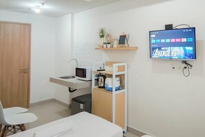 A compact hotel room with a kitchenette, a TV mounted on the wall, a wooden door, and a small seating area. - Neat And Warm Studio Room At Apartment Bandaraya - Tallasa City Makassar (Makassar)