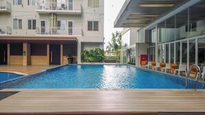 A swimming pool with a wooden deck in the foreground, surrounded by a modern building with balconies and large windows. - Comfortable Studio (No Kitchen) At Bogor Icon Apartment (Bogor)