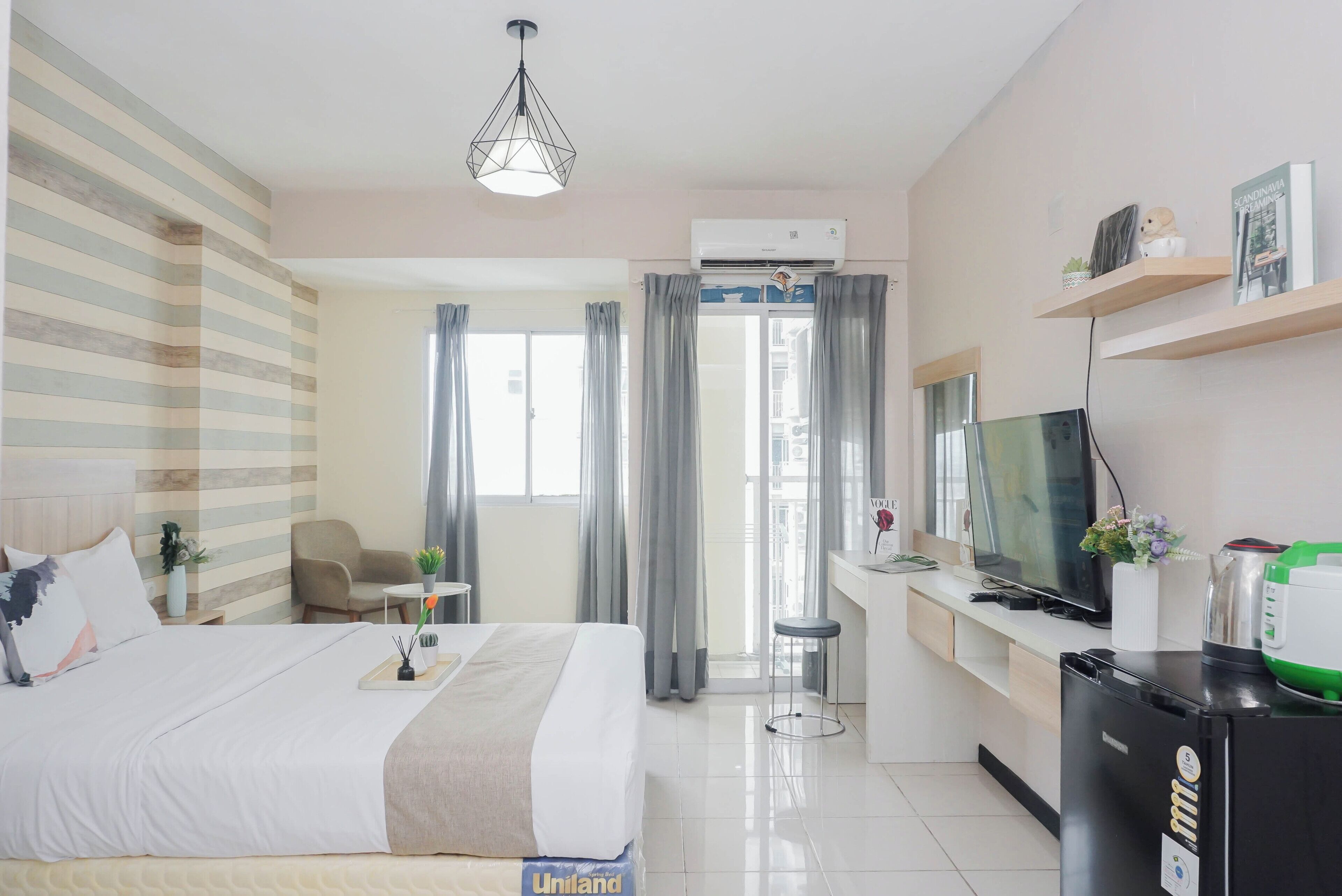 A hotel room with a bed, bedside table, TV, refrigerator, and a desk with a chair.
