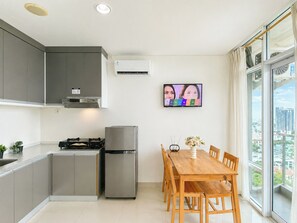 A modern kitchen with grey cabinets, a built-in stove, a fridge, and a dining area with a wooden table and chairs. - Homey Studio Room At Apartment Brooklyn Alam Sutera (South Tangerang)