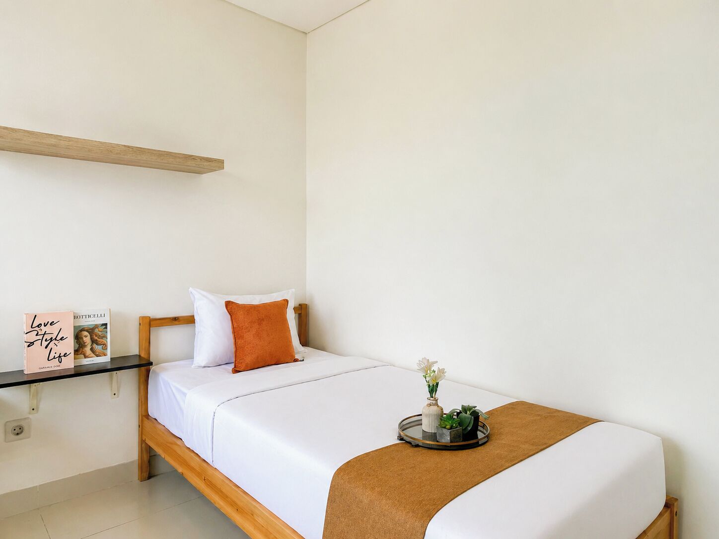 A single bed with white bedding, an orange pillow, a small bedside table with a vase of flowers, and a shelf with a book and a sign.