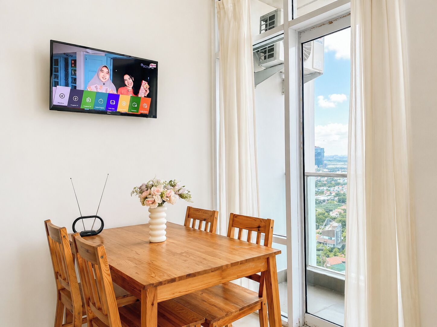 A dining area with a wooden table, chairs, a vase with flowers, and a mounted television displaying a program.