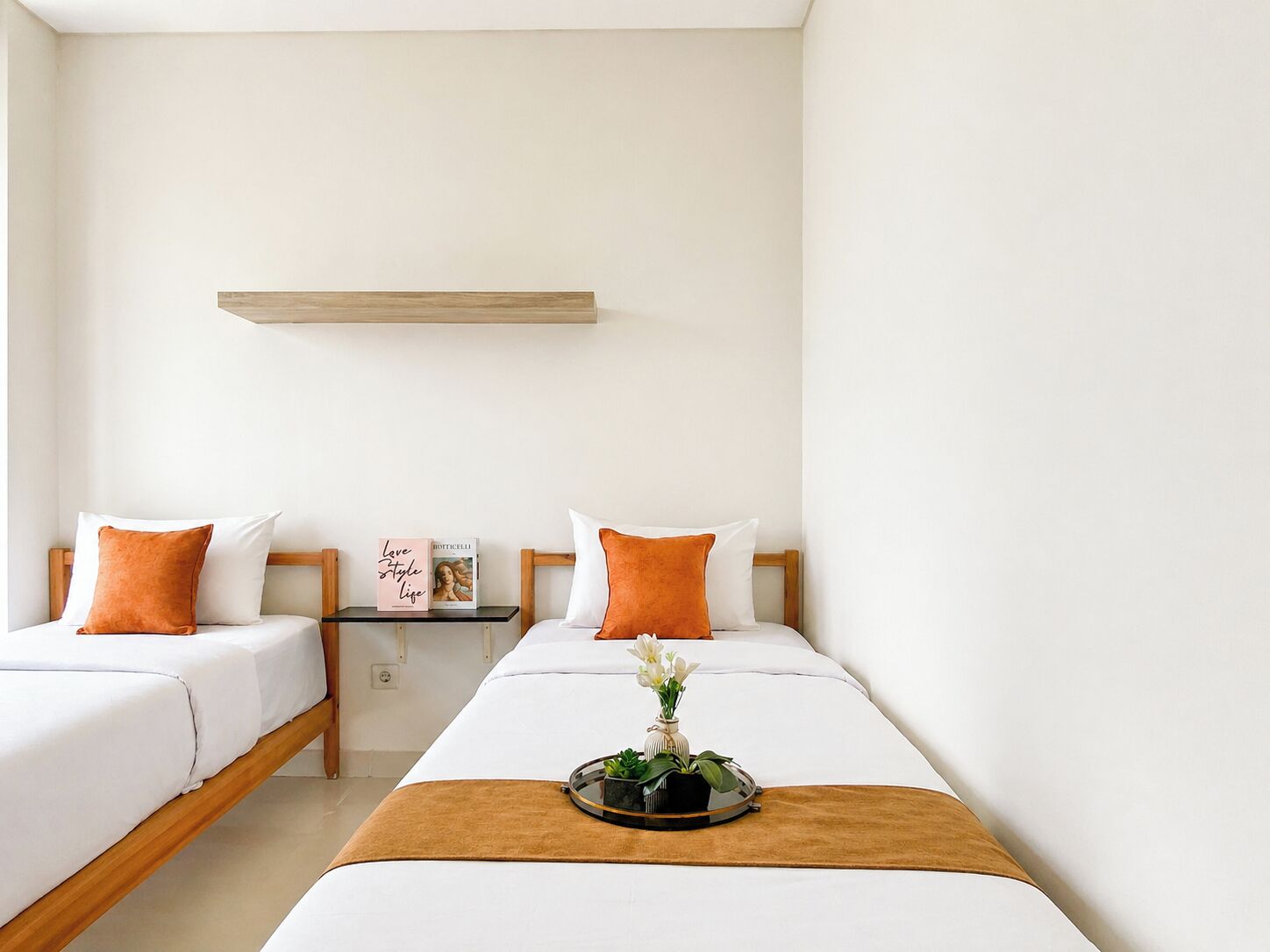 Habitación de hotel con dos camas, una mesita de noche con un libro, un estante pequeño sobre la cama y una bandeja con flores en el suelo.