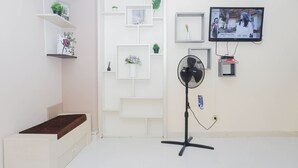 Una habitación con cama, ventilador, televisor y estanterías con plantas y libros.
