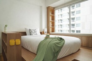 A modern bedroom with a large bed, a wooden wardrobe, a window with a view of a residential building, and a bedside table with a lamp. - 1 Pax Wooden Touch Studio At Citra Living Apartment (Jakarta)