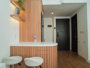 A modern kitchen with a wooden accent wall, a white countertop, and two bar stools. - 1 Pax Wooden Touch Studio At Citra Living Apartment (Jakarta)