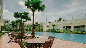 Zona junto a la piscina con mesas y sillas de madera, rodeada de palmeras y una piscina.