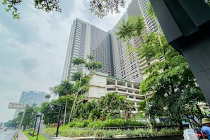 A modern high-rise building with a mix of glass and concrete facades, surrounded by lush greenery and palm trees. - Stylish 1Br At Collins Boulevard Apartment (Tangerang)