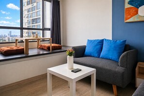 Ein modernes Wohnzimmer mit einem grauen Sofa, blauen Kissen, einem weißen Tisch mit einer Vase und einem Smartphone, und Blick auf die Stadt durch große Fenster.