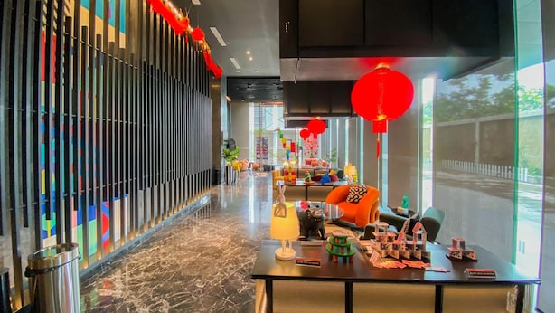 Um lobby de hotel moderno com um corredor longo, painéis verticais coloridos e uma área de estar com uma mesa exibindo maquetes de casas.