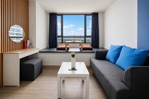 Un salon moderne avec un canapé, une table basse, une petite table ornée d’un vase, et une vue sur la ville grâce à de grandes fenêtres.