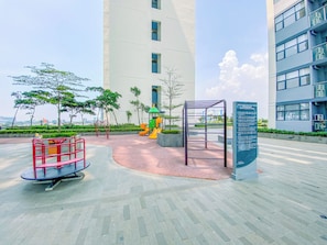Ein Spielplatz mit Rutschen und Klettergerüsten, umgeben von einem befestigten Bereich mit Bänken und einem Hinweisschild.