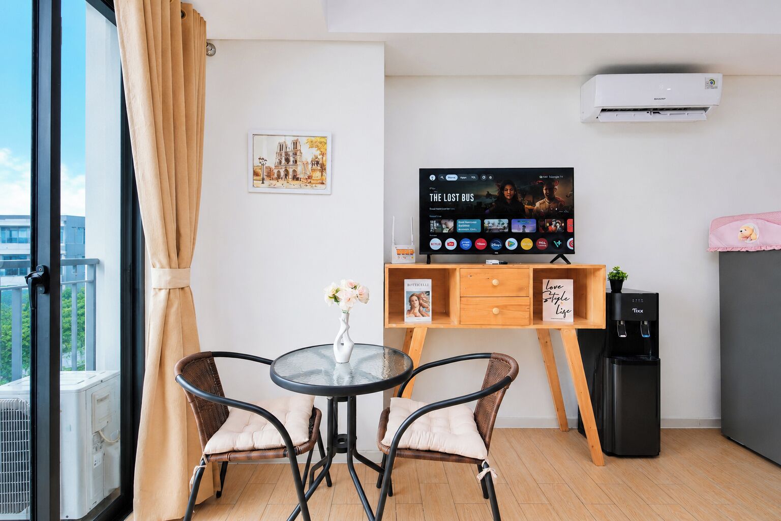 A modern room with a glass table, two cushioned chairs, a wooden desk with a TV, a water dispenser, and an air conditioner.